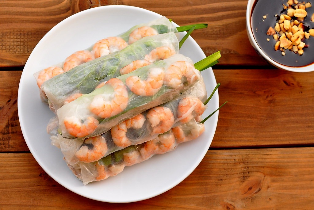 An overhead view of a plate of beautifully arranged Goi Cuon spring rolls, garnished with fresh herbs and served with a side of peanut dipping sauce, on a rustic wooden table.